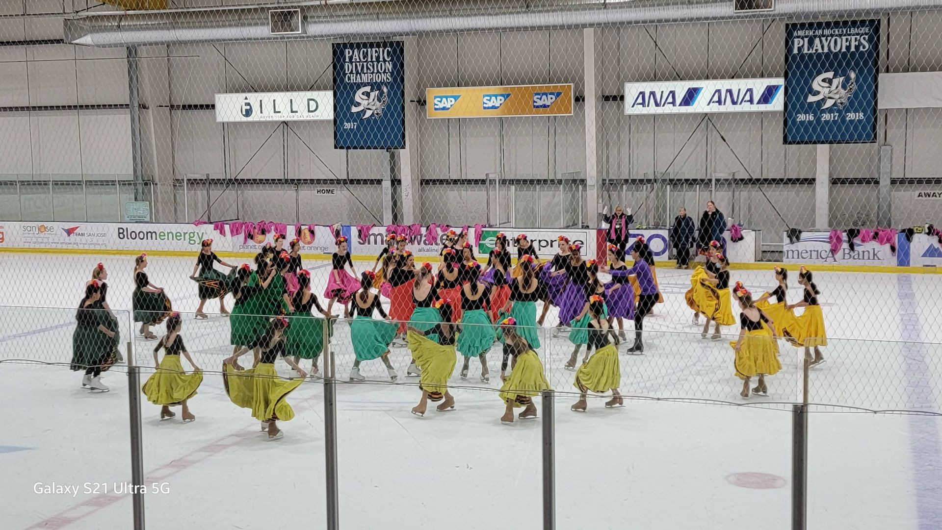 Group of performers in colorful costumes ice skating inside a rink with banners and ads on the walls.